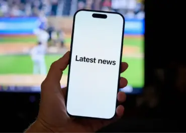 Main tenant un smartphone affichant le texte « Latest news » à l’écran, avec un match de baseball flou en arrière-plan sur un écran de télévision.